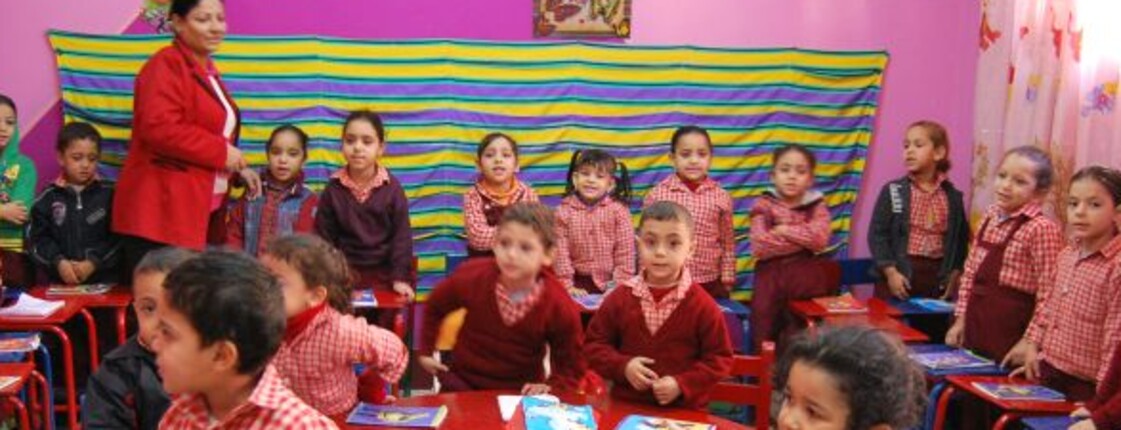 Blick in einen Kindergartenraum: die Wände sind rosa und geschmückt, die Kinder stehen hinter kleinen Pulten.