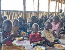 Kinder sitzen am Boden und essen aus Tellern, die vor ihnen stehen