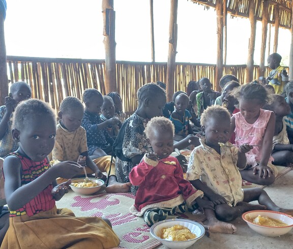 Kinder sitzen am Boden und essen aus Tellern, die vor ihnen stehen