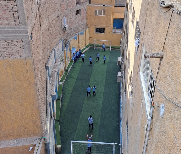 Blick von oben auf den Fußballplatz einer ägyptischen Grundschule, wo gerade Mädchen Fußball spielen. Der Platz ist zwischen hohen Häusern eingebettet.