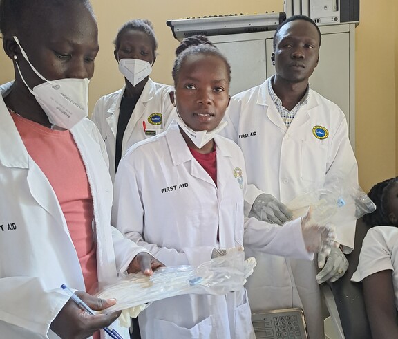 Drei junge Südsudanes*innen stehen in Laborkitteln und Mundschutz ein einem Raum. Die vorderste Person hält ein Klippboard in der Hand.