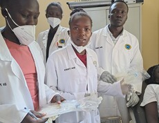 Drei junge Südsudanes*innen stehen in Laborkitteln und Mundschutz ein einem Raum. Die vorderste Person hält ein Klippboard in der Hand.